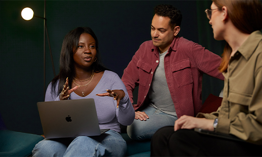 Ginelle hat ein MacBook auf dem Schoß und arbeitet mit zwei Kolleg:innen zusammen.
