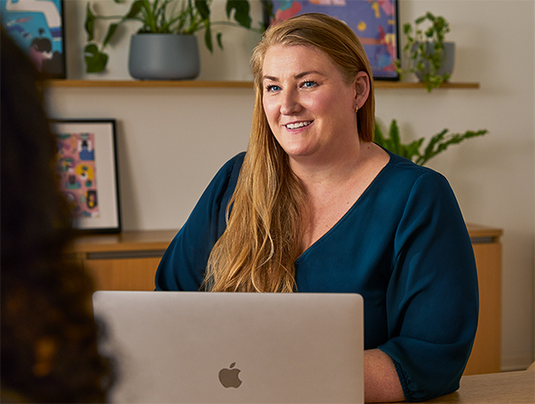 Una empleada de Apple sentada en la mesa de una zona común utiliza un MacBook mientras habla con alguien de su equipo.