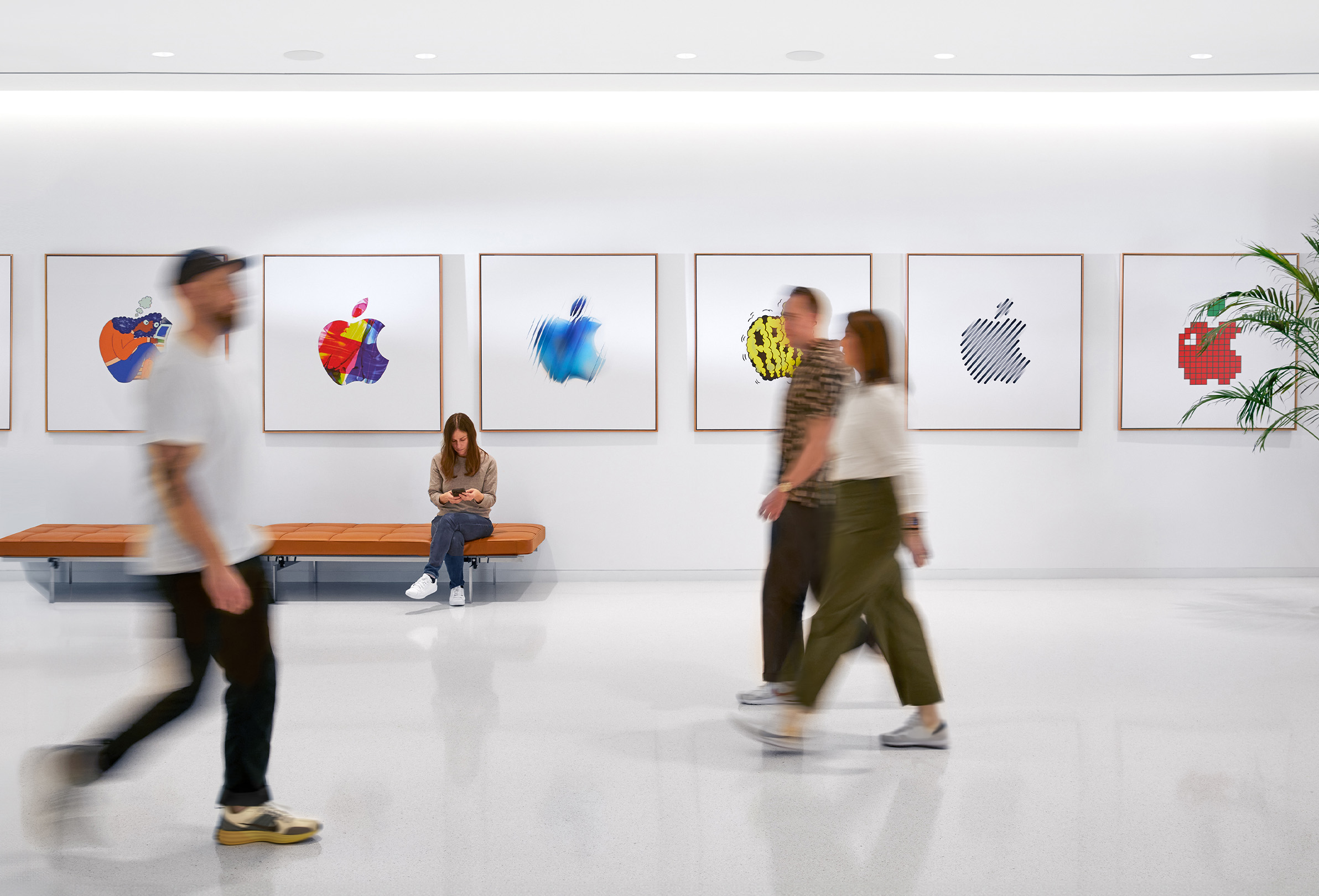 Três colegas passam por uma parede com logotipos da Apple coloridos e uma quarta funcionária está sentada em um banco.