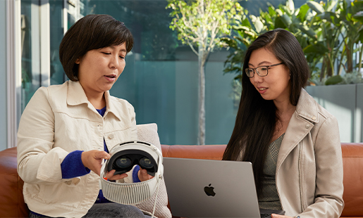 Jessica utiliza un MacBook en un espacio interior con acceso al patio mientras una compañera de Apple le enseña el Apple Vision Pro.