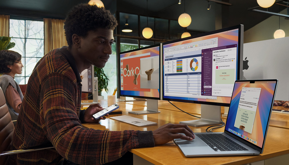Una persona in un ufficio usa una mano per digitare su un MacBook Air aperto collegato a due monitor esterni e a una Magic Keyboard, mentre con l’altra tiene un iPhone