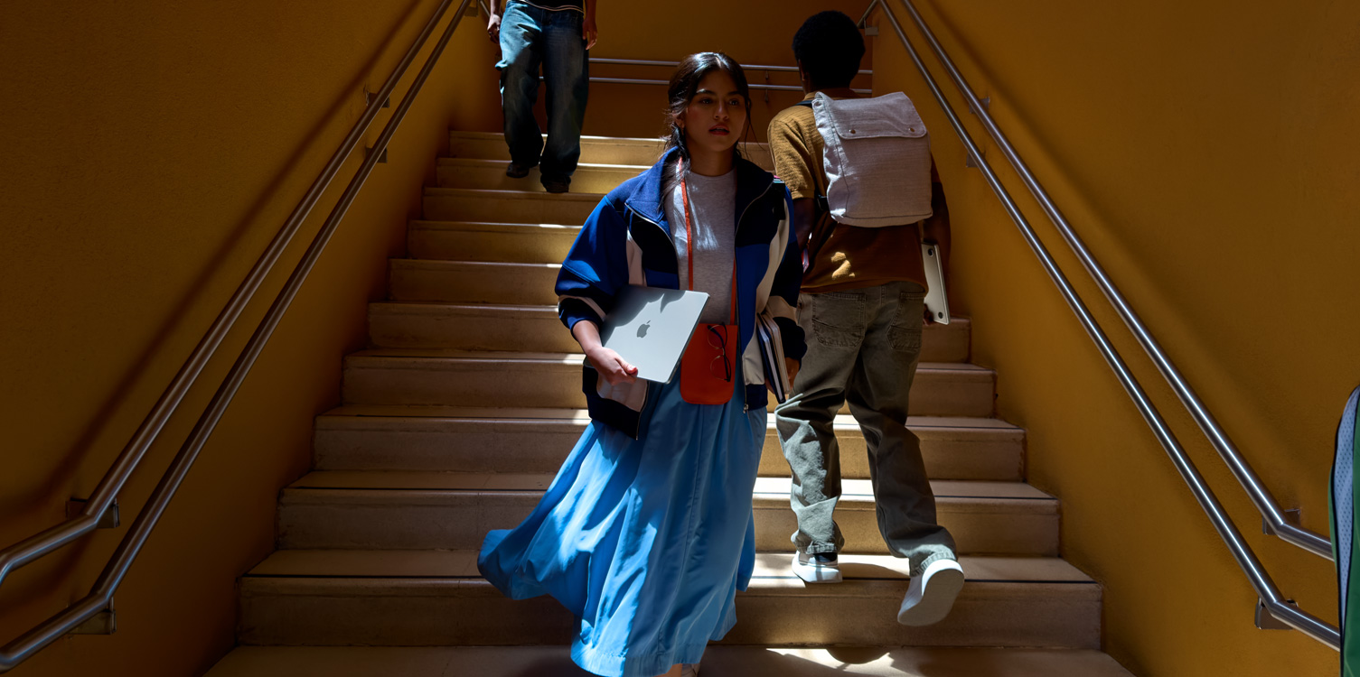 Une étudiante descend un escalier. Elle tient un MacBook et des livres.