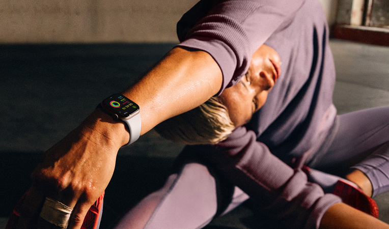 Una donna sul pavimento che fa stretching allungando il braccio verso la gamba mentre indossa un Apple Watch Series 10, il display mostra Allenamento, Defaticamento, Attività, Anelli Attività