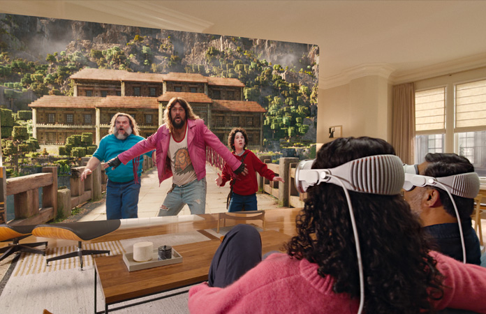 Apple Vision Pro를 착용한 두 사람이 소파에 나란히 앉아 방 전체에 펼쳐진 화면에 표시된 마인크래프트 무비를 보고 있는 모습